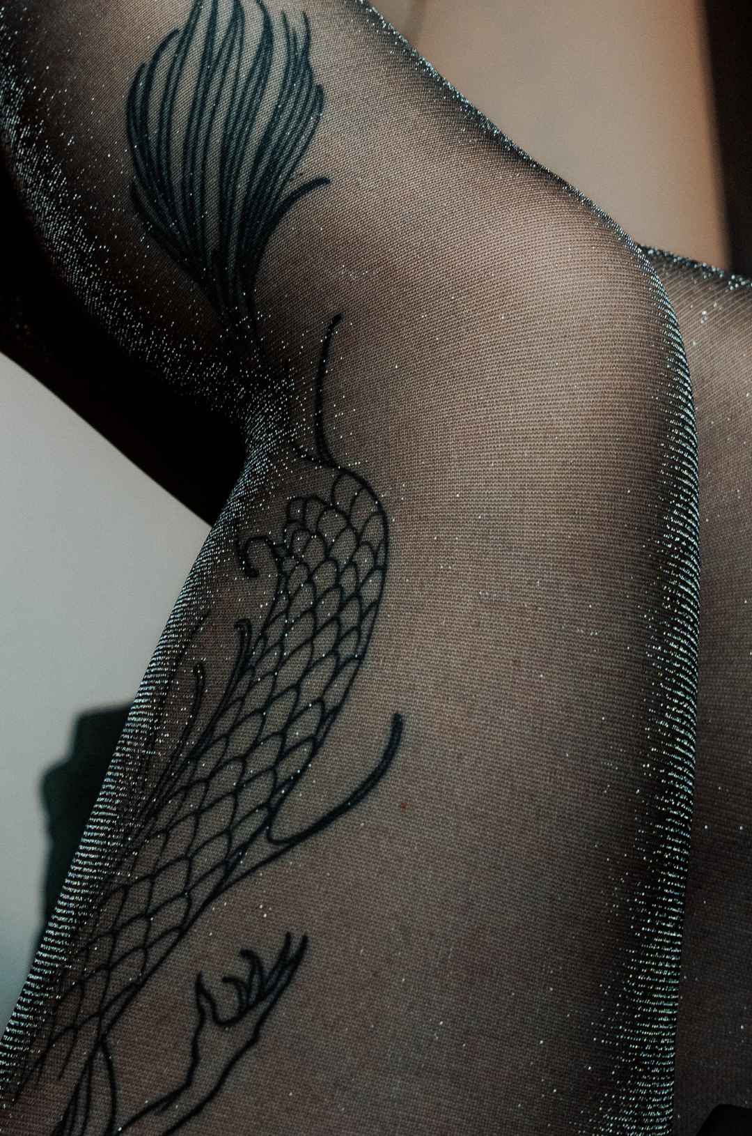 Detailaufnahme des silbern glitzernden Stoffs der Glitzerstrumpfhose. Durch den Stoff der Feinstrumpfhose scheint das Tattoo des Models durch.